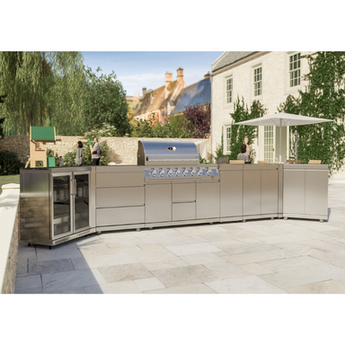 A luxury outdoor kitchen featuring a stainless steel 6-burner gas BBQ, sink with tap, and fridge set on a marble-effect surface, surrounded by a white house and garden in the background.