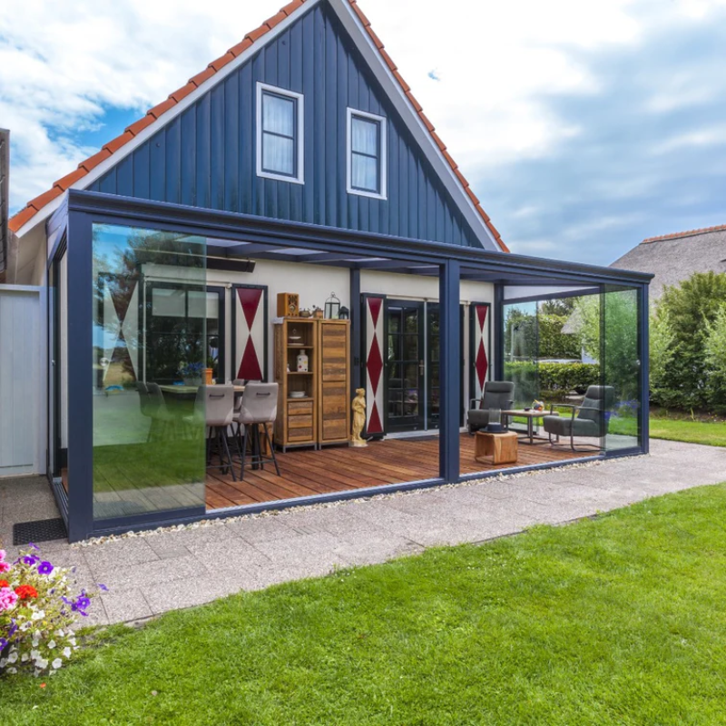 Aluminium veranda with clear polycarbonate roof, featuring a dark frame and posts, attached to a blue house with a thatched roof.