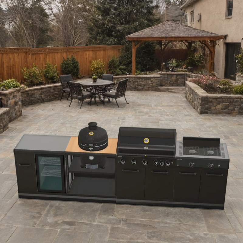Modular outdoor kitchen made of stainless steel with a 5-burner gas barbecue, stove, fridge, and a kamado cabinet, set on an outdoor patio with a table and chairs in the background.