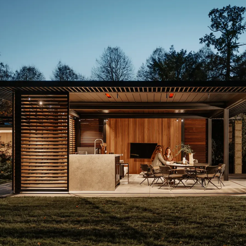 Modern outdoor kitchen and dining area with people sitting at a table, surrounded by trees and a Renson Amani Pergola